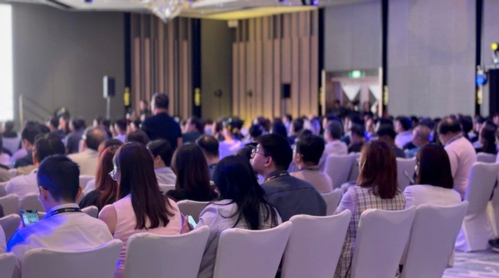 A large audience sitting in a conference hall, facing a presentation stage, with a mix of attendees in business attire.