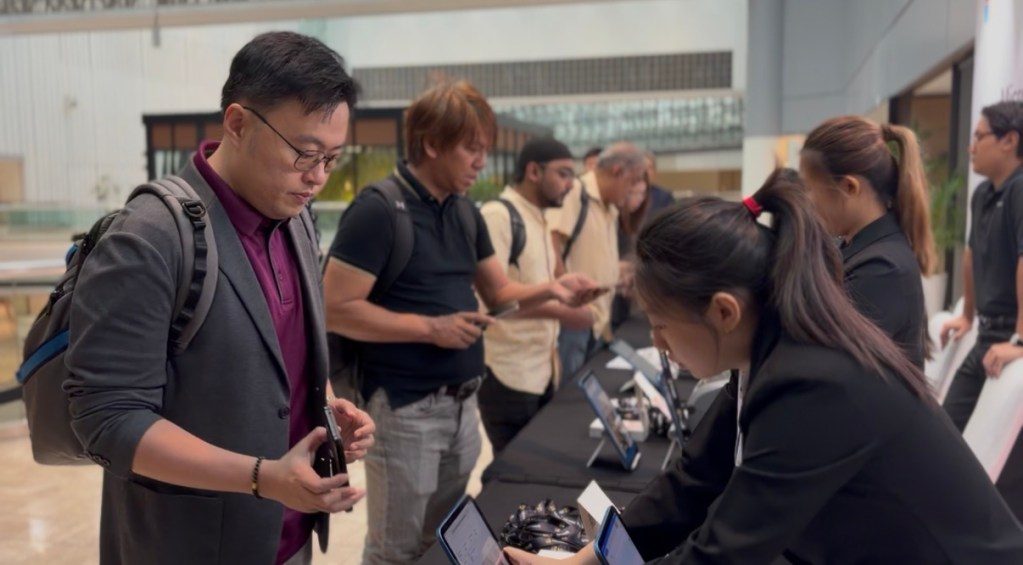 A group of people engaged in a technology-related event, with individuals examining devices at a display table. One man in formal attire is looking closely at a device, while others interact with staff and check their phones.