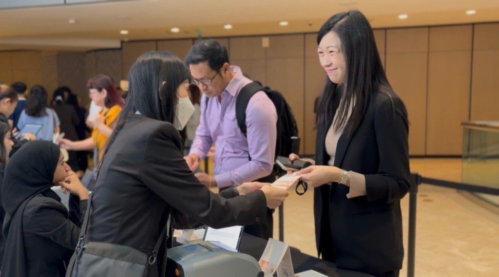 A professional woman in a black suit is handing a card to another woman wearing a mask at a registration desk, while a third person in a light purple shirt observes in the background.