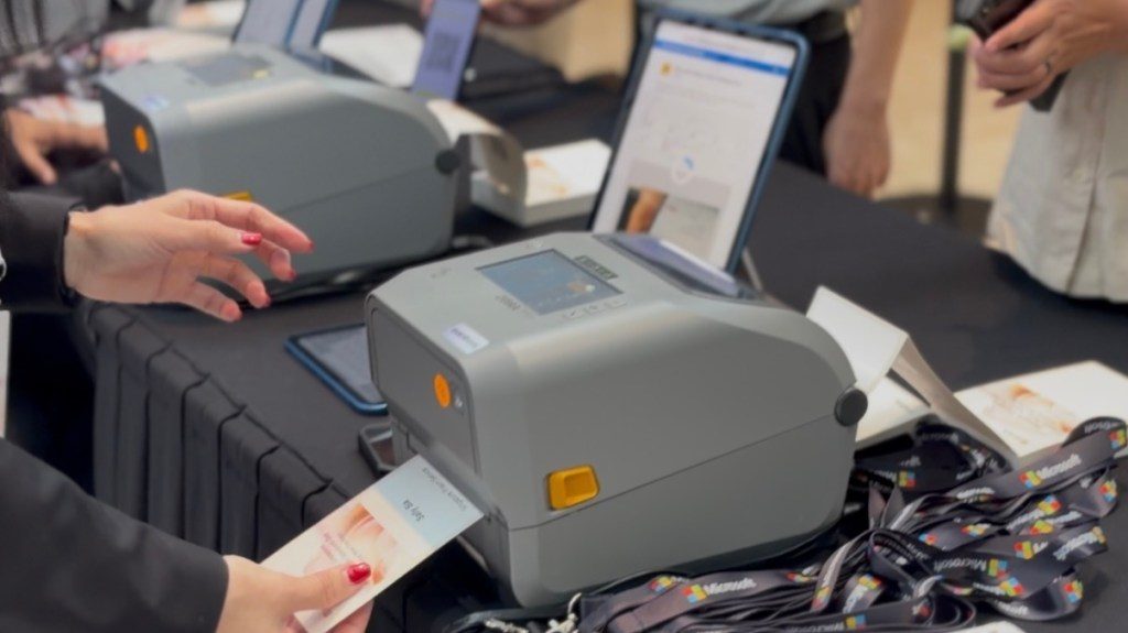 A hand with red nails operates a card printer at a display table, with multiple printers and digital devices in the background.